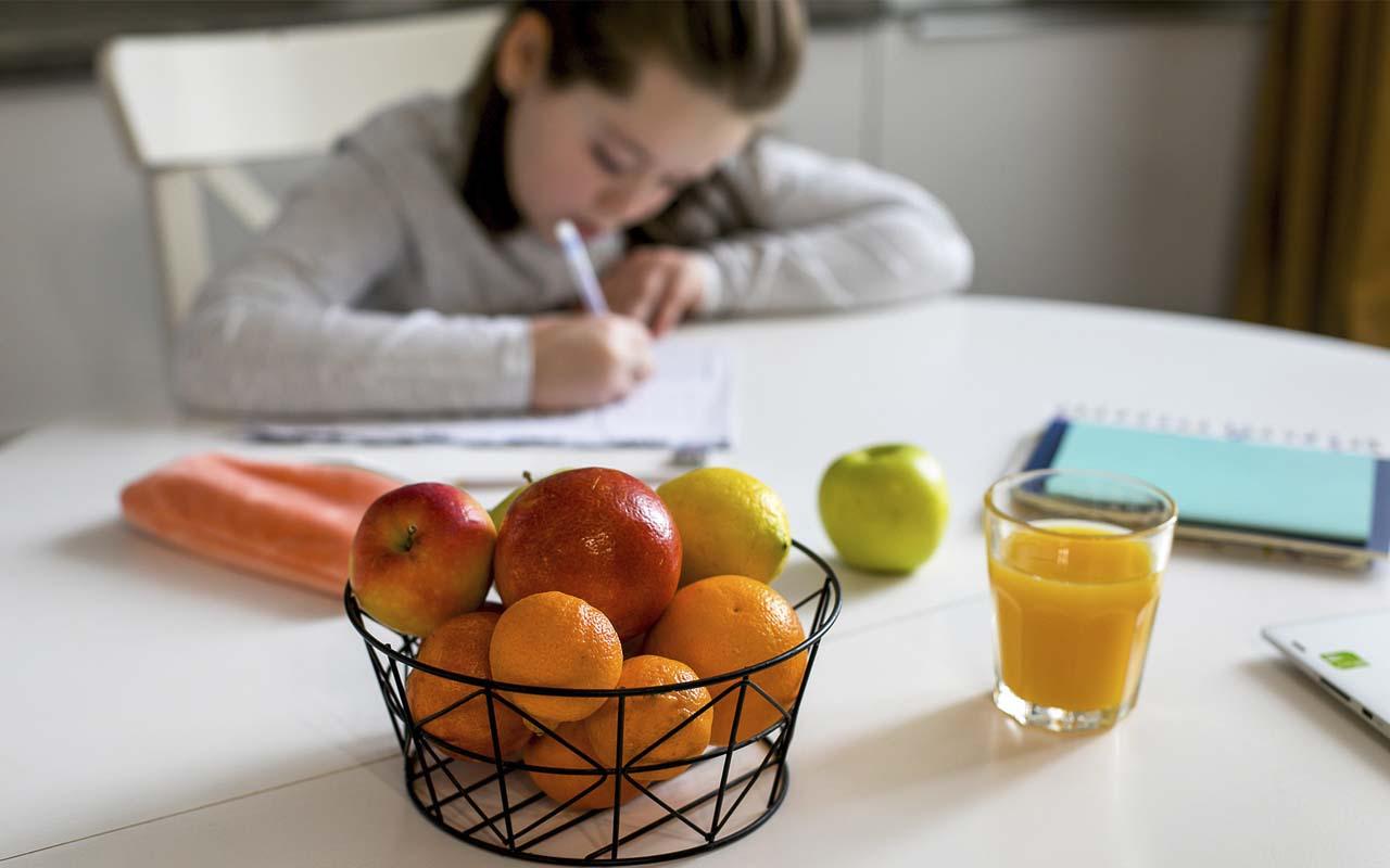 Young girl doing her homework at the kitchen table with orange juice and a bowl of fruit in focus