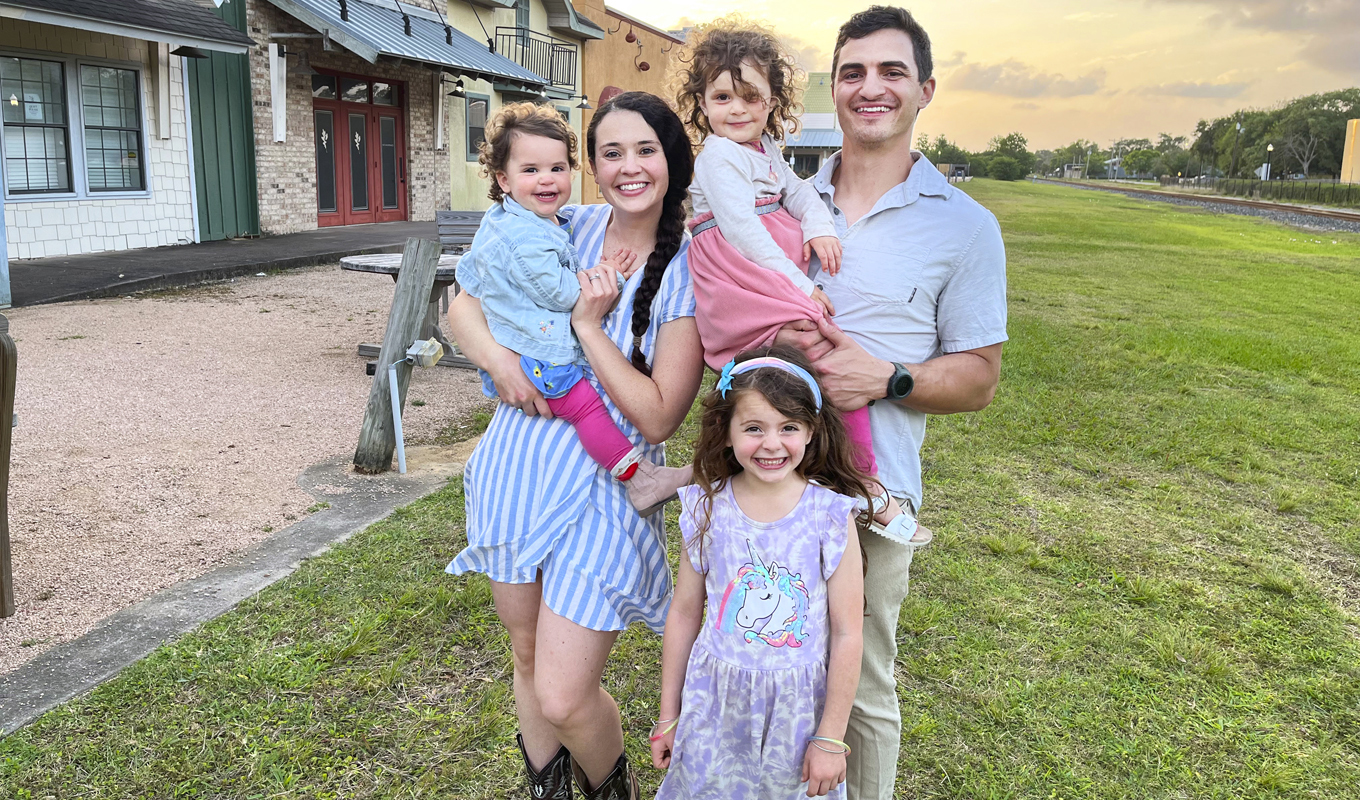 Family of five poses outdoors. Smiling parents hold two young daughters, while older daughter stands in front, grinning.