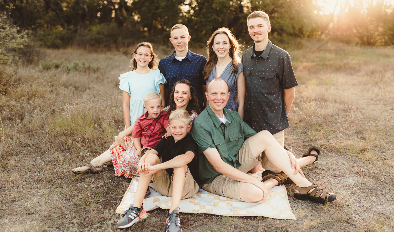 A family of eight poses on a grassy field; some are seated on a blanket, others stand behind.