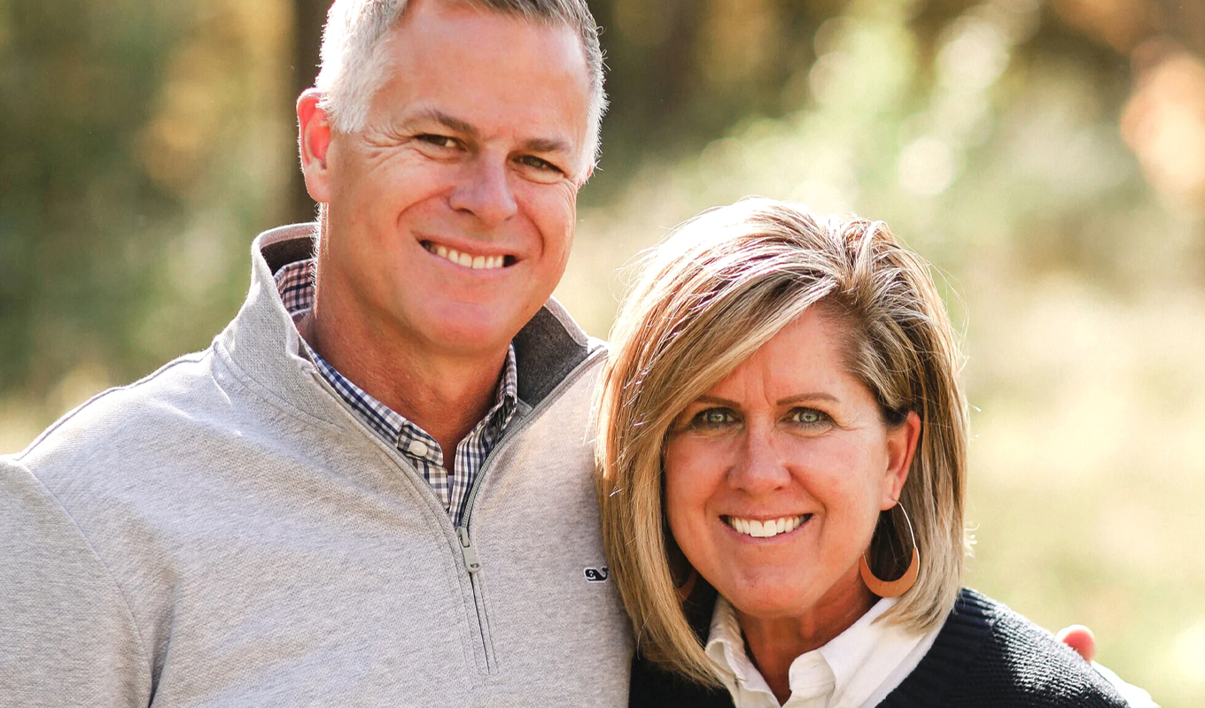 Couple standing outdoors with arms around each other, dressed in sweaters, with trees and greenery in the background.