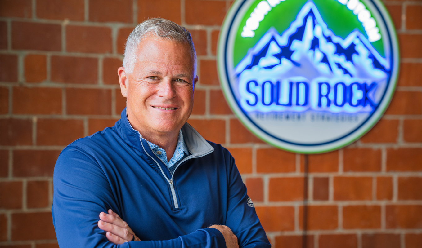 Person in blue shirt stands with arms crossed in front of brick wall and "SOLID ROCK" adventure-themed sign.