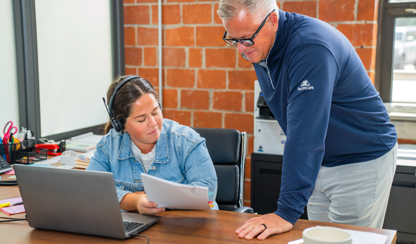 wo people in an office; one seated with headphones and a document, the other standing and looking at it, with a brick wall in the background.