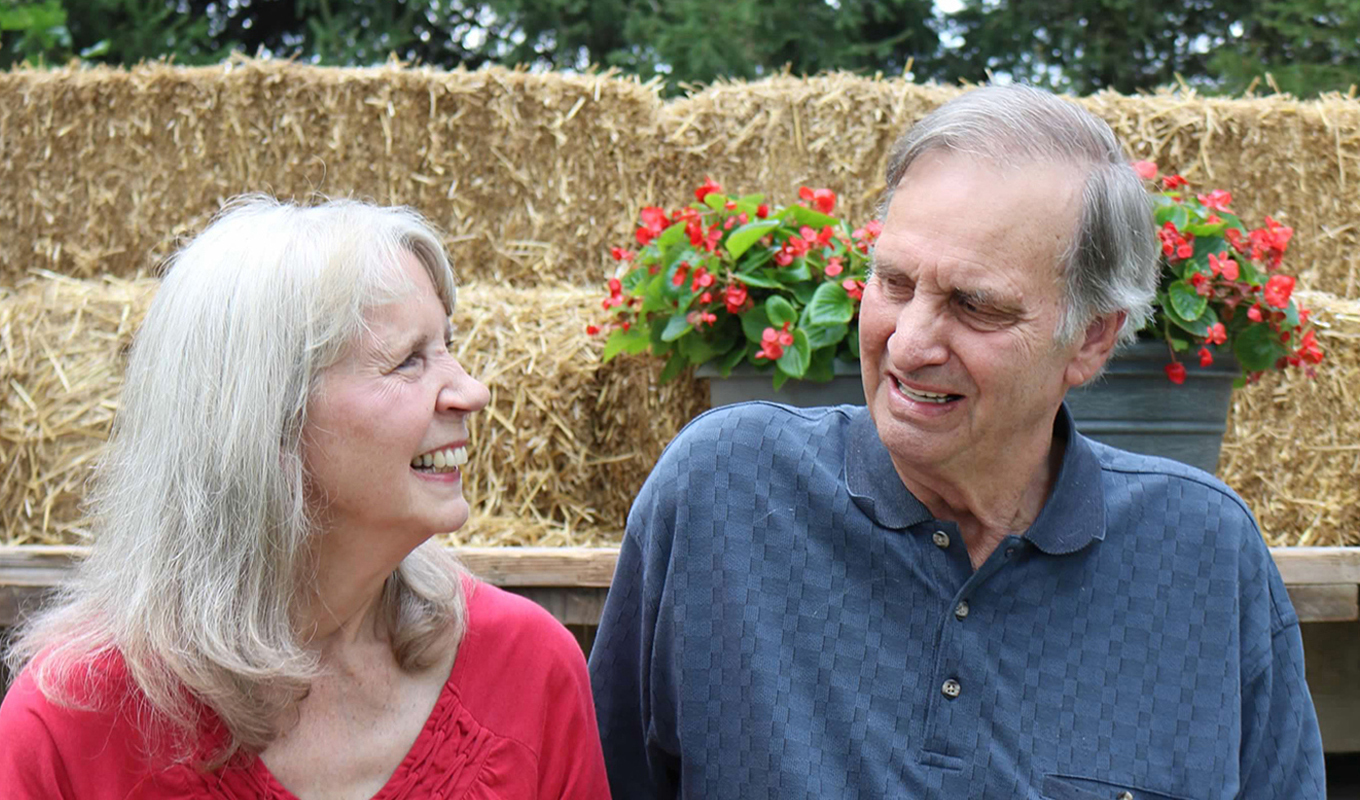 Two elderly individuals laughing and enjoying a conversation outdoors, seated in front of a backdrop of hay bales and red flowers.