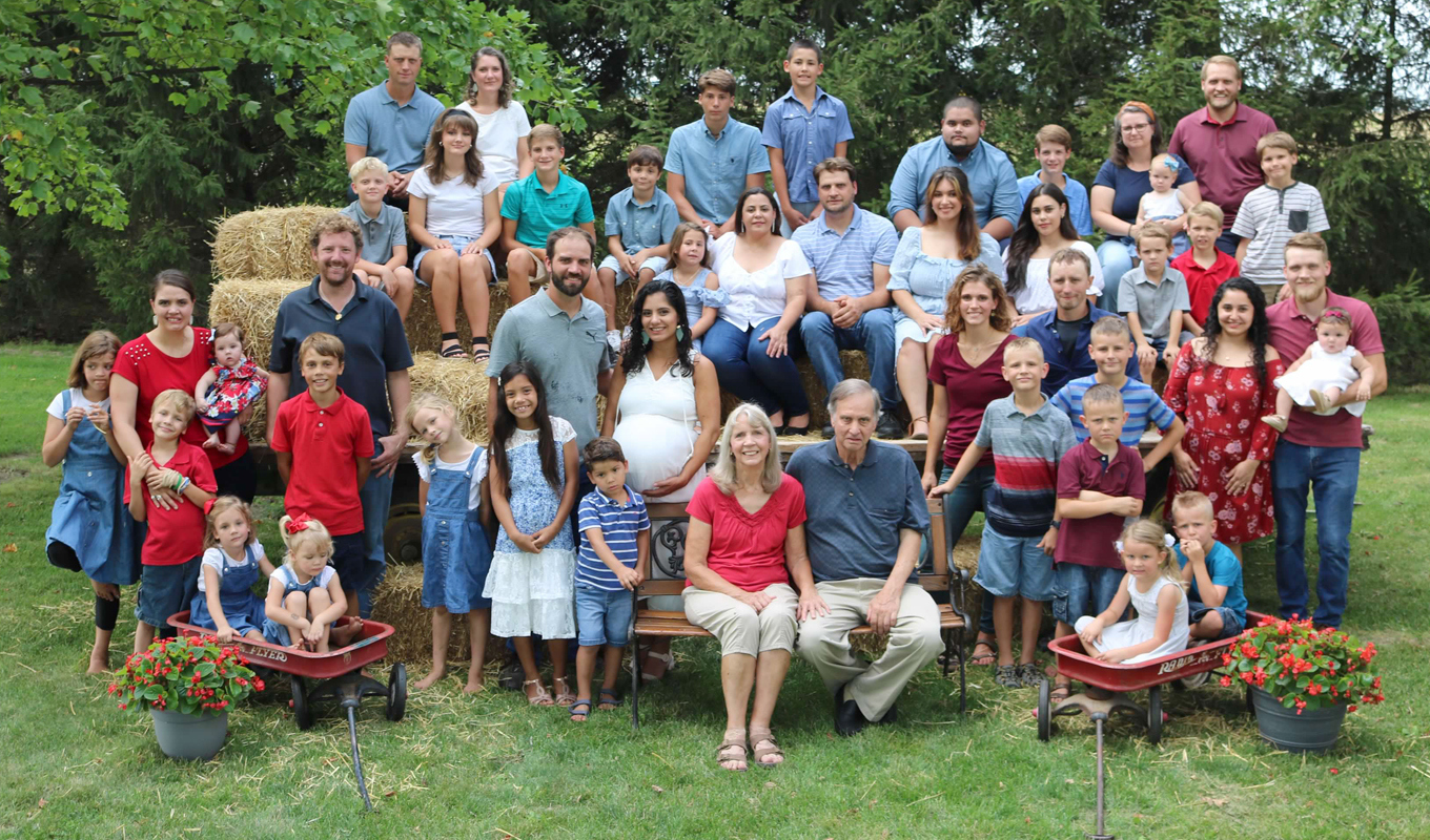 Large family group of 39 people outdoors, seated on hay bales and benches, surrounded by trees and greenery.