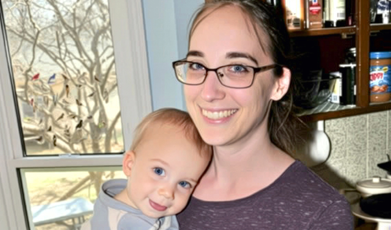 Smiling woman with glasses holding a resting child in a cozy kitchen with shelves and a decorated window in the background.