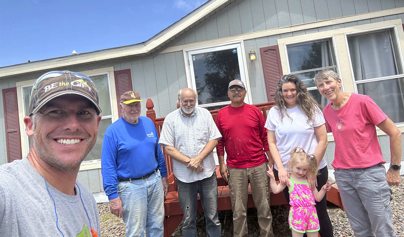 Group of seven people smiling in front of a house with red shutters and a porch.