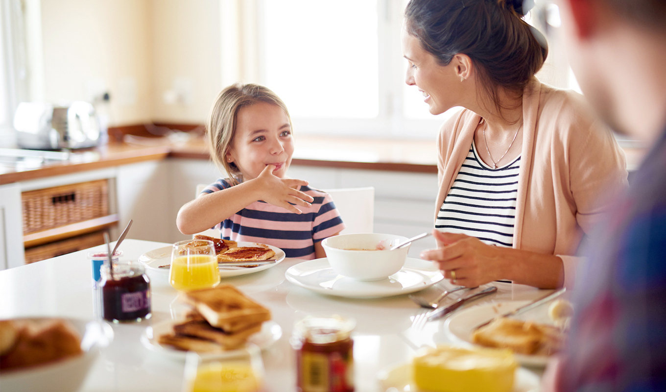 A child and an adult sit at a kitchen table set with toast, jam, orange juice, and cereal, enjoying breakfast in a bright kitchen with natural light.