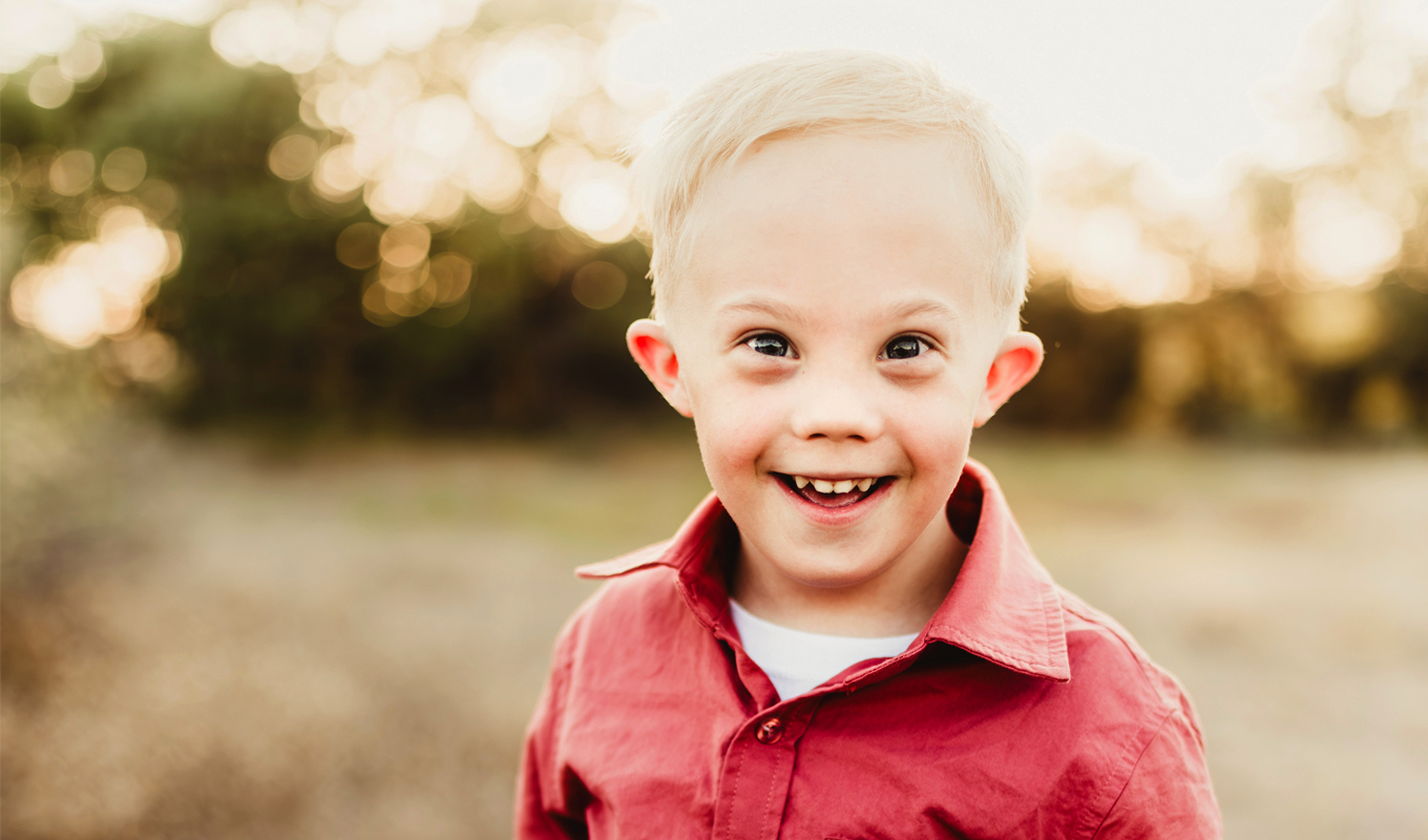 A child in a red shirt and beige shorts stands in a sunlit field with dry grass and trees.