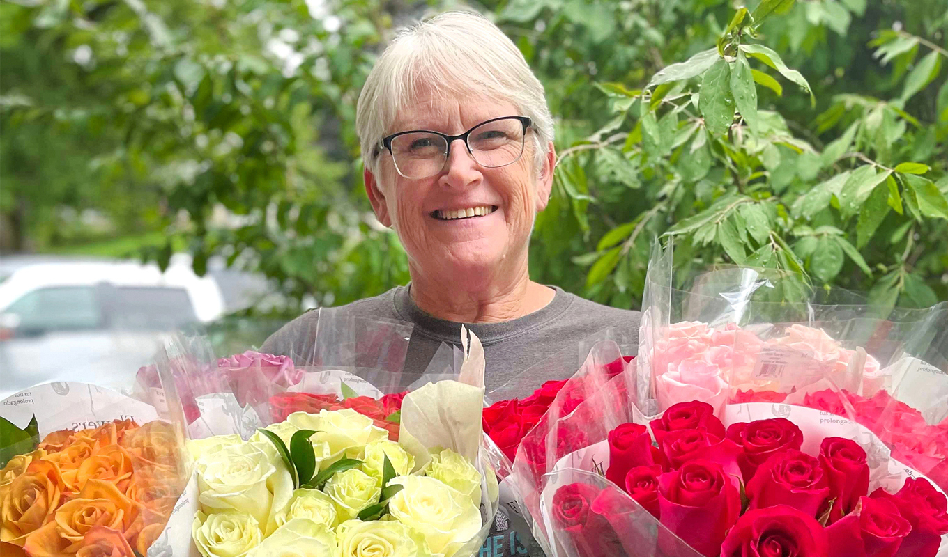 A person holds three large bouquets of roses—orange, white, and red—wrapped in clear plastic and placed in black containers, standing outdoors with greenery and a parked car in the background.