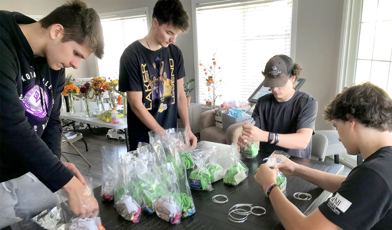 Four people are seated around a table, assembling items into clear plastic bags. The table holds supplies such as scissors and rubber bands. The room is well-lit with large windows and floral decorations in the background.