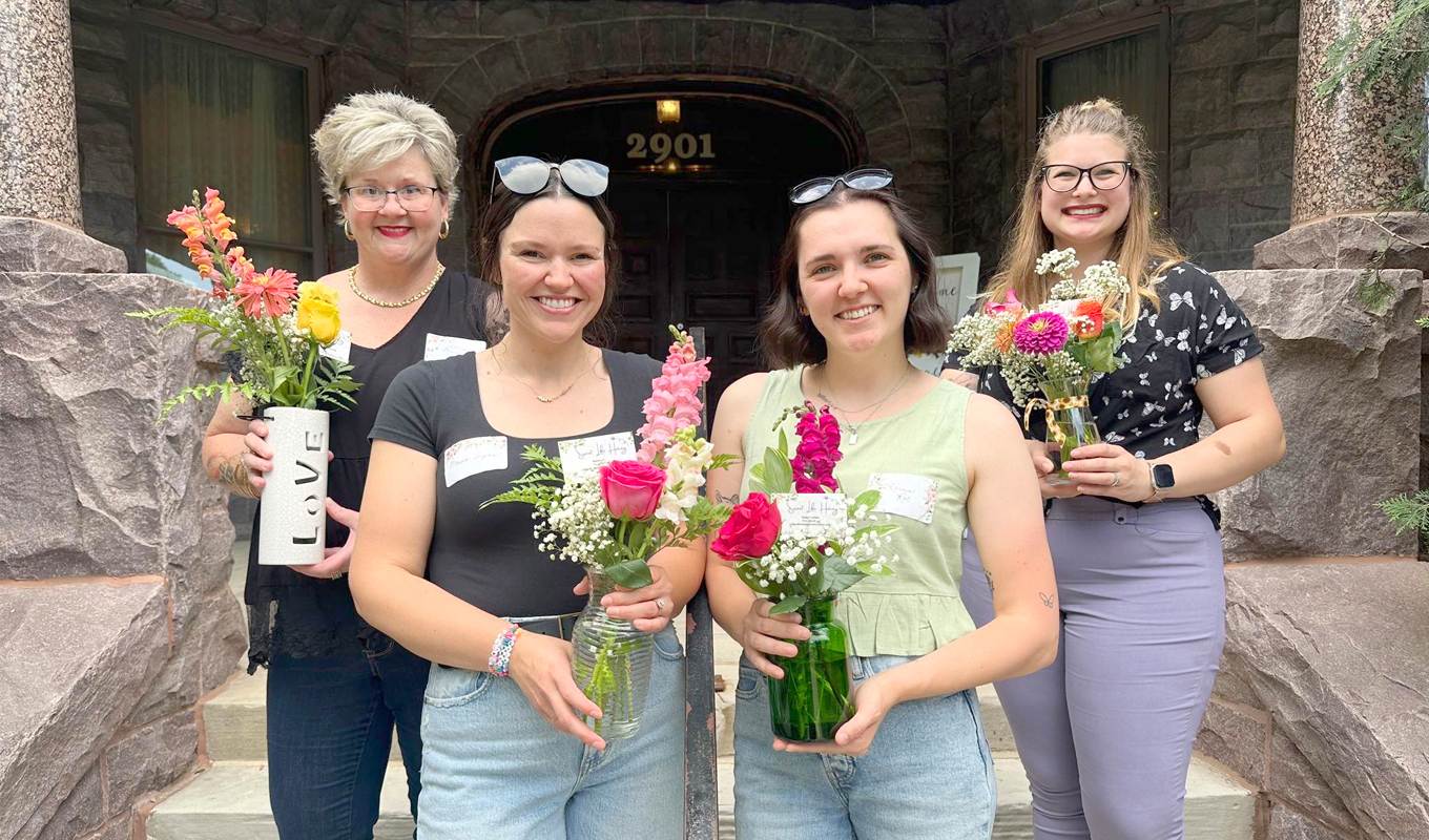 Four individuals stand on stone steps in front of a building marked '2901,' each holding a vase or jar of colorful flowers, including roses. They are dressed casually. The background shows a stone facade and a wooden door.