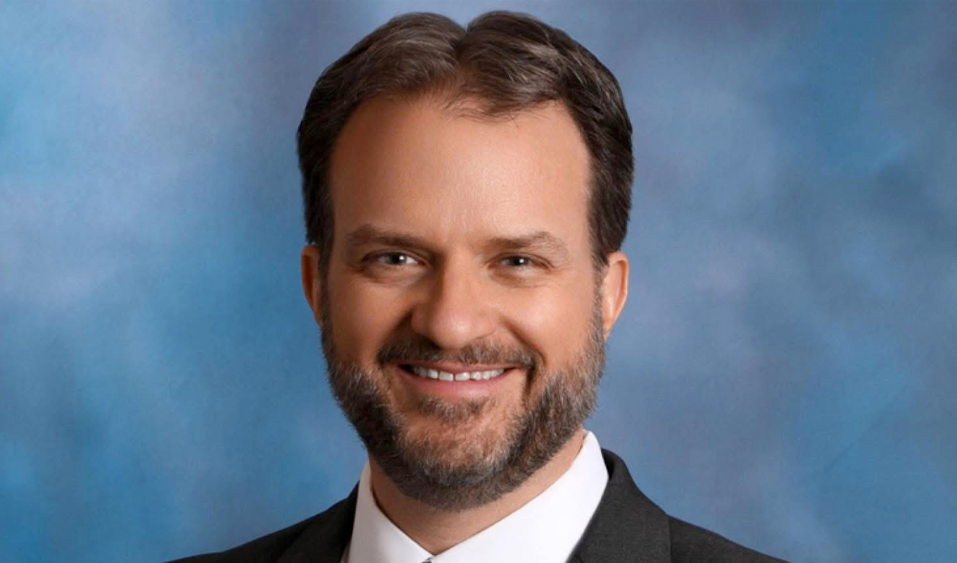 Professional headshot of a person with short dark hair and a beard, wearing a suit and tie, against a plain blue background.