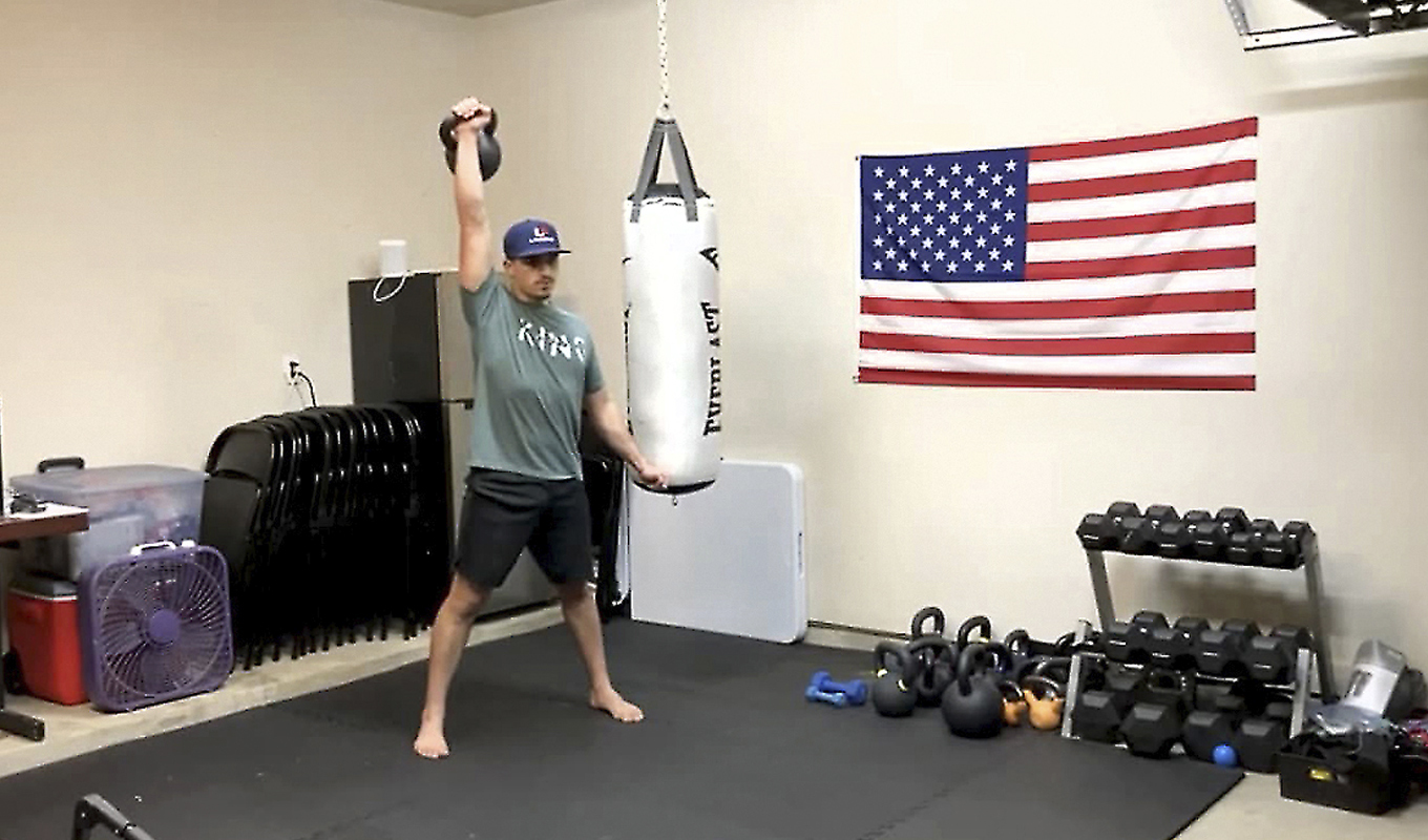 A man in a home gym presses a kettlebell over his head.