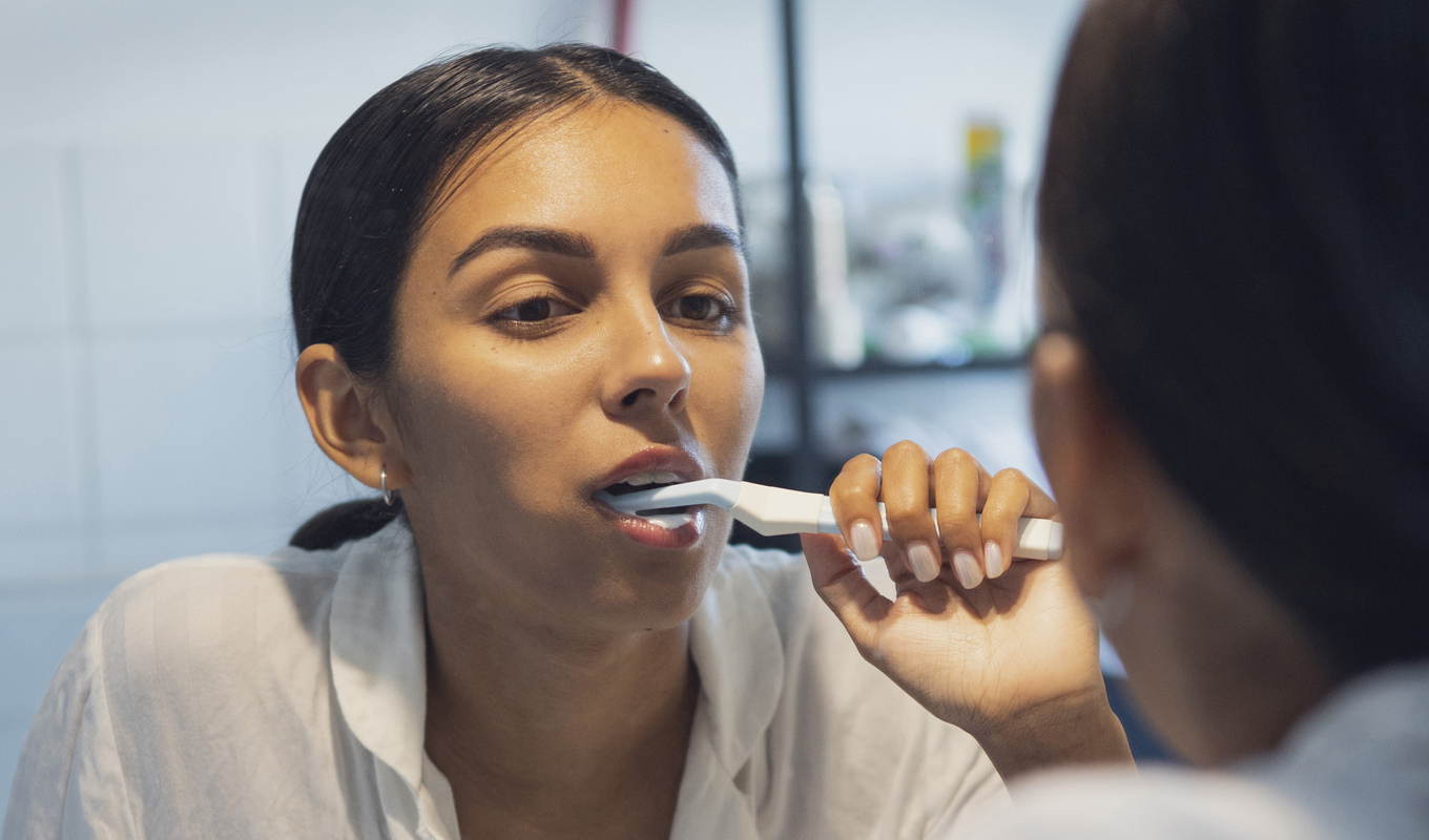A woman stands in front of a mirror, brushing her teeth with a focused expression on her face.