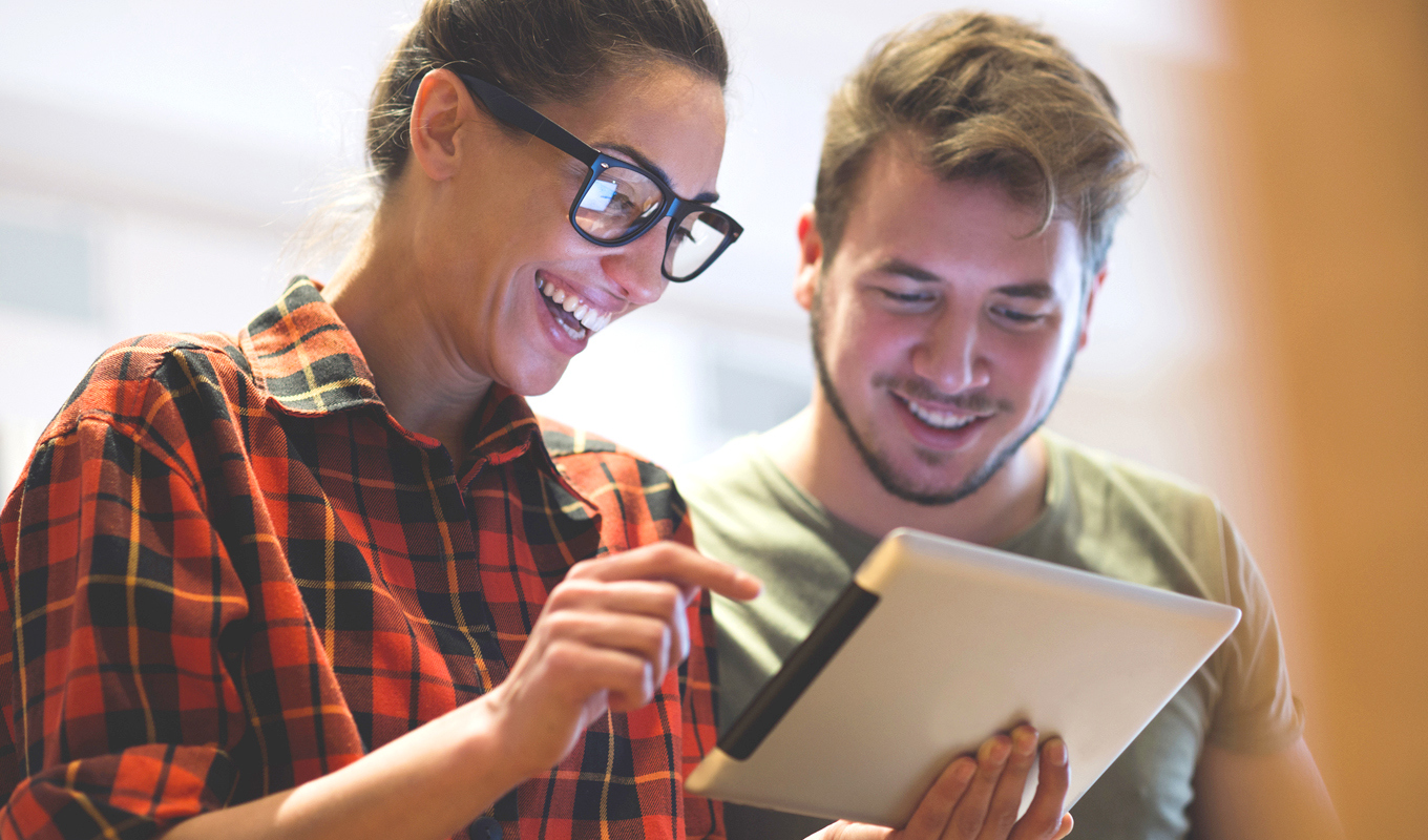 Couple smiling and looking at a tablet. Woman in a plaid shirt points at the screen.