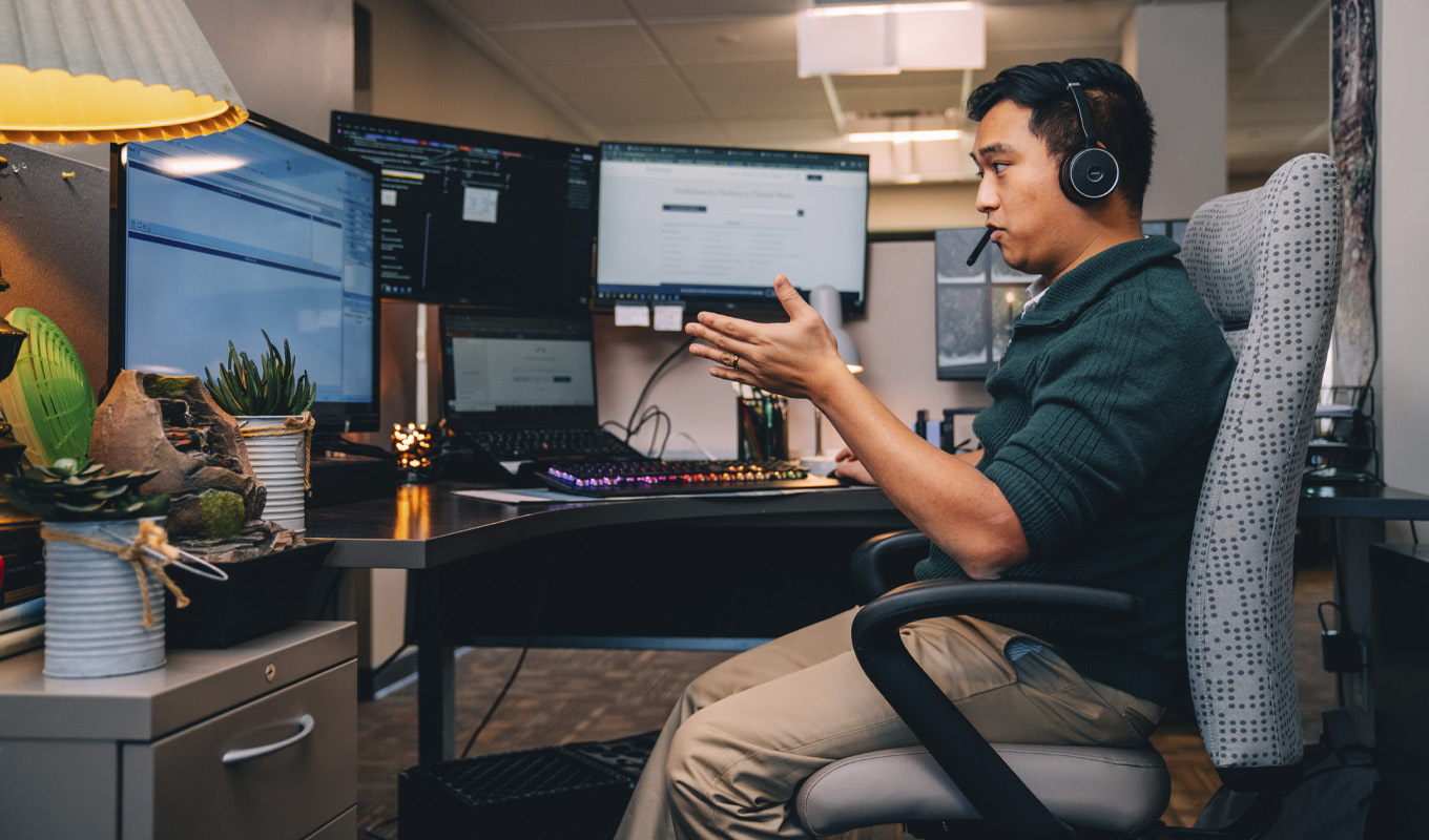 A man seated at a desk, working on two monitors and a laptop, focused on his tasks in a professional environment.