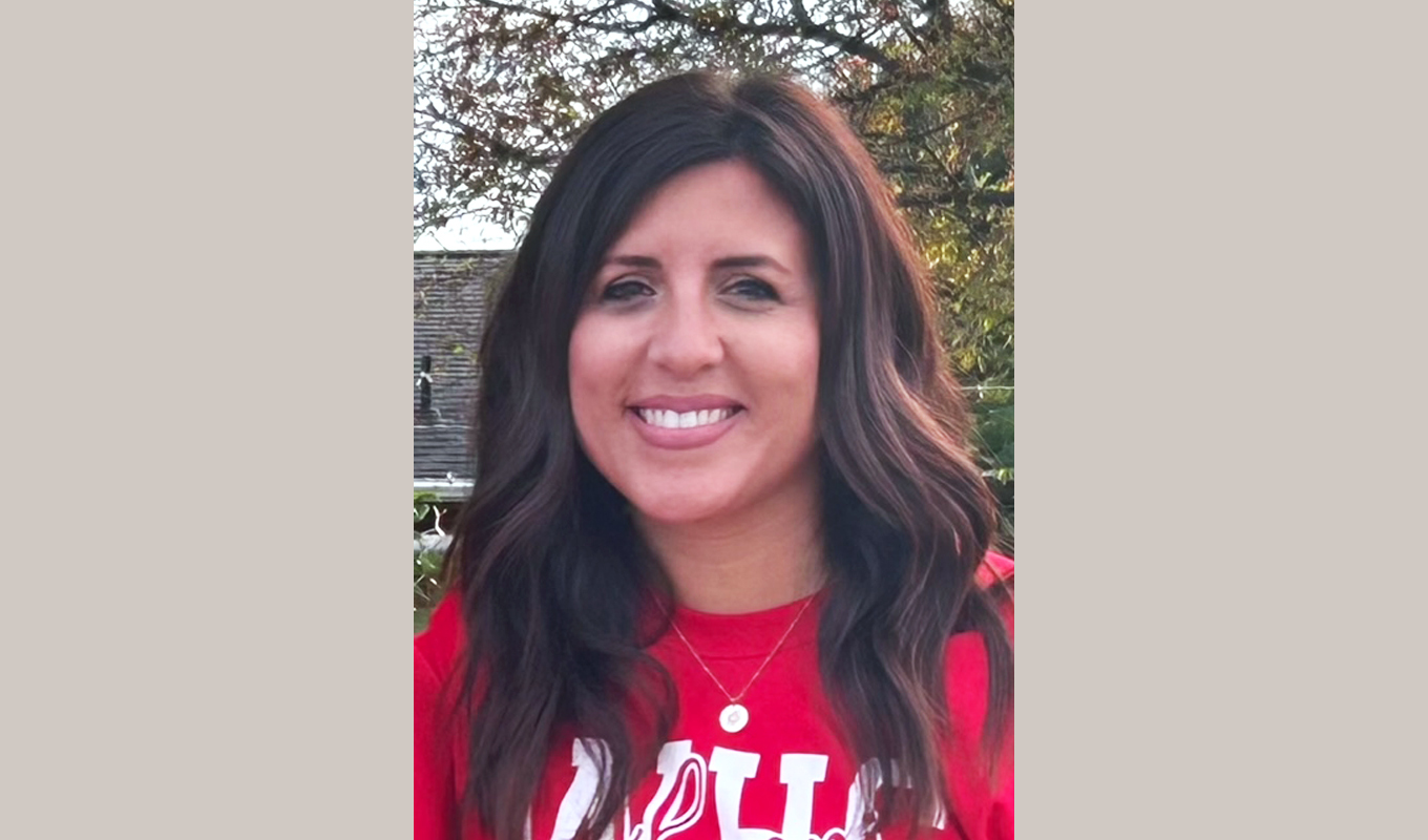 A person with long, wavy dark hair wearing a red shirt with white lettering and a necklace with a round pendant. The background includes trees and part of a building.