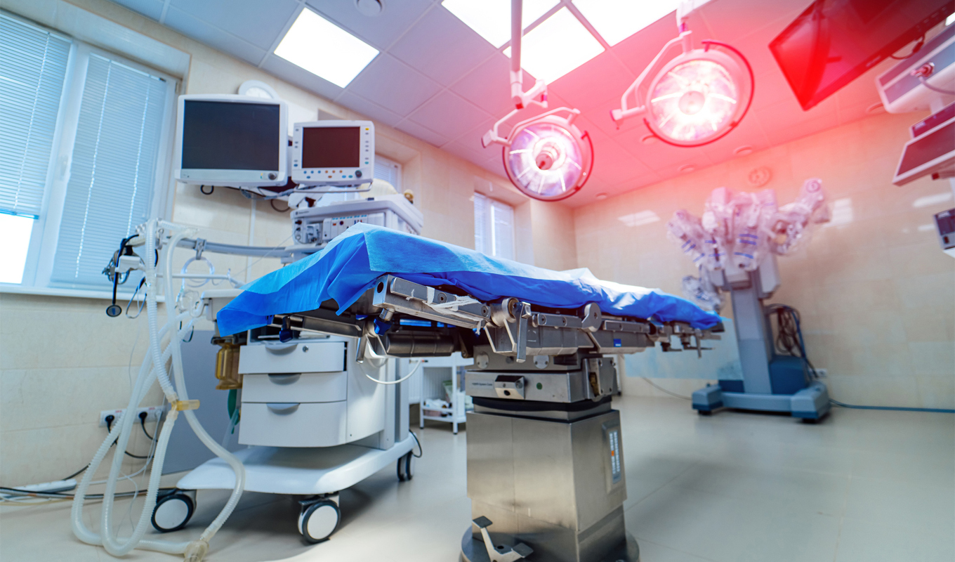 A modern operating room with a surgical table covered in blue sterile drapes at the center, surrounded by medical equipment including anesthesia machines, monitors, and articulated robotic surgery arms. Overhead surgical lights cast a bright red glow, and large windows with blinds line the walls.