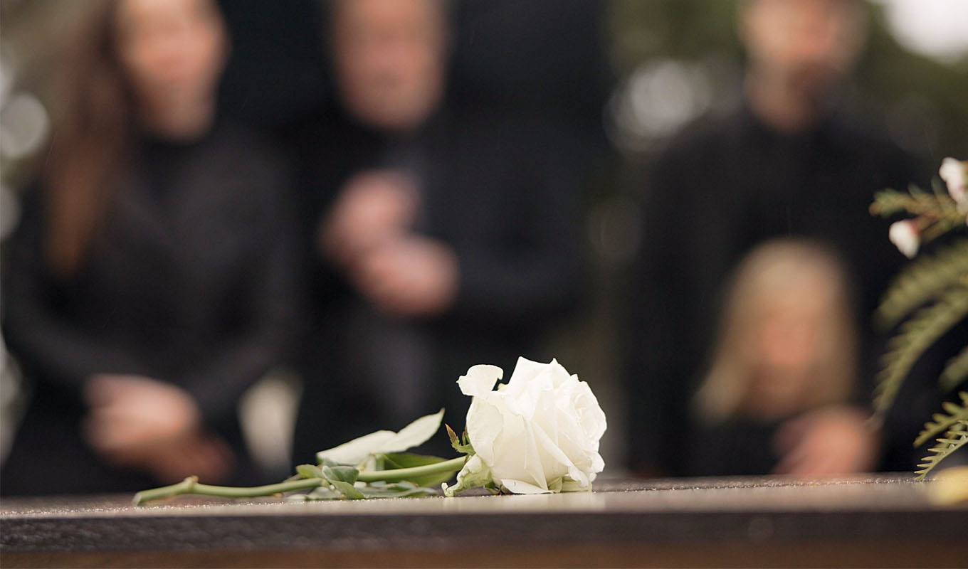 A white rose rests on a wooden casket, with several people standing nearby in soft focus.