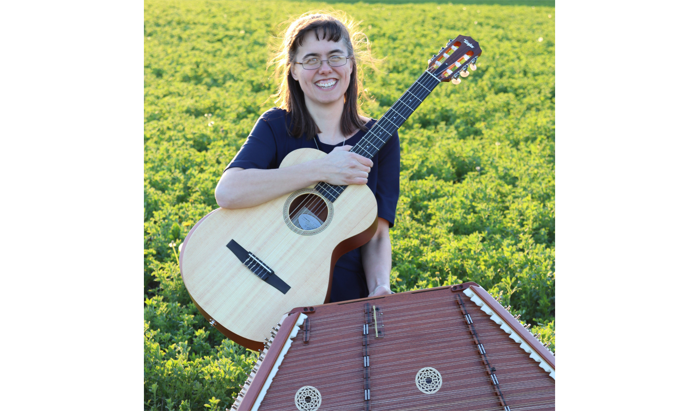 A woman stands in a field near a hammered dulcimer, holding an acoustic guitar, surrounded by tall grass and a clear blue sky
