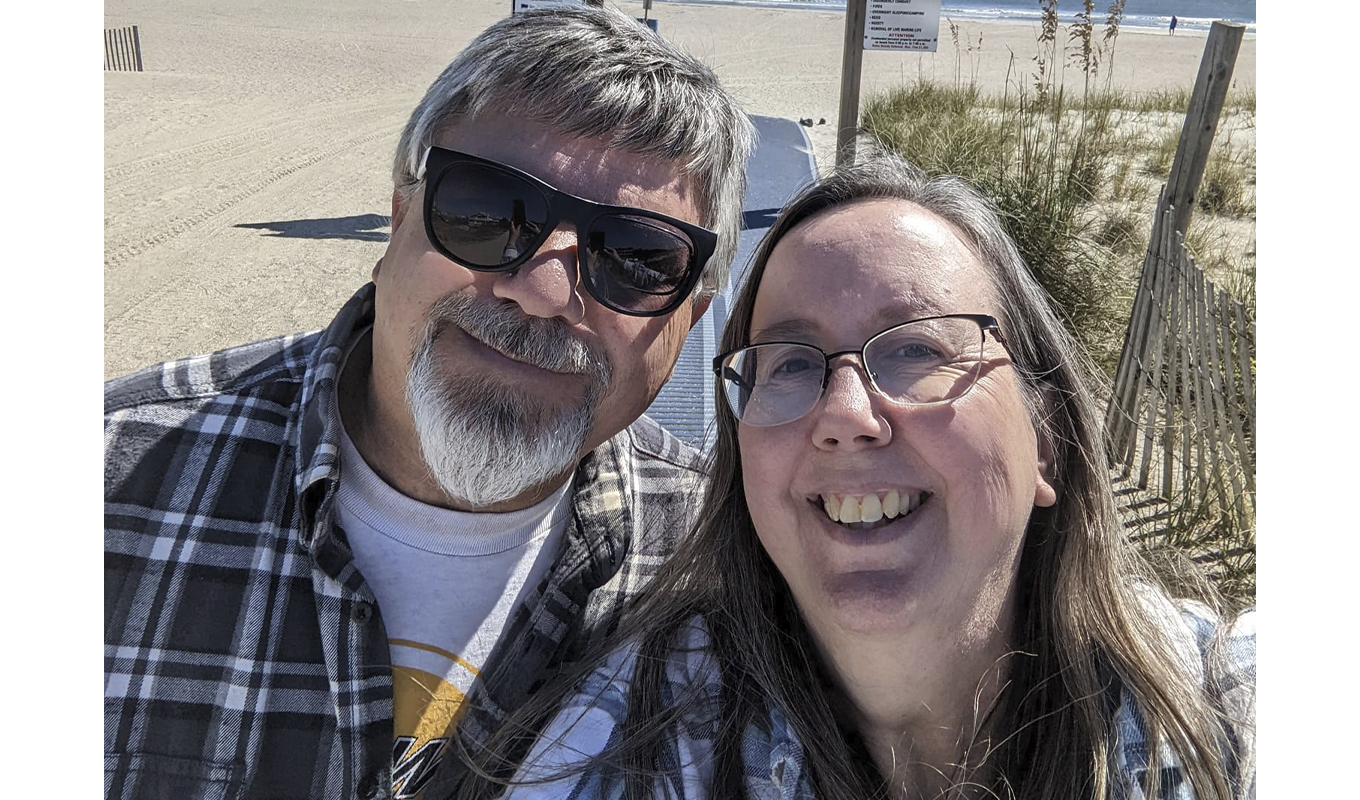 A husband and wife smiling as they take a selfie together on a sunny beach, with waves gently crashing in the backgrou