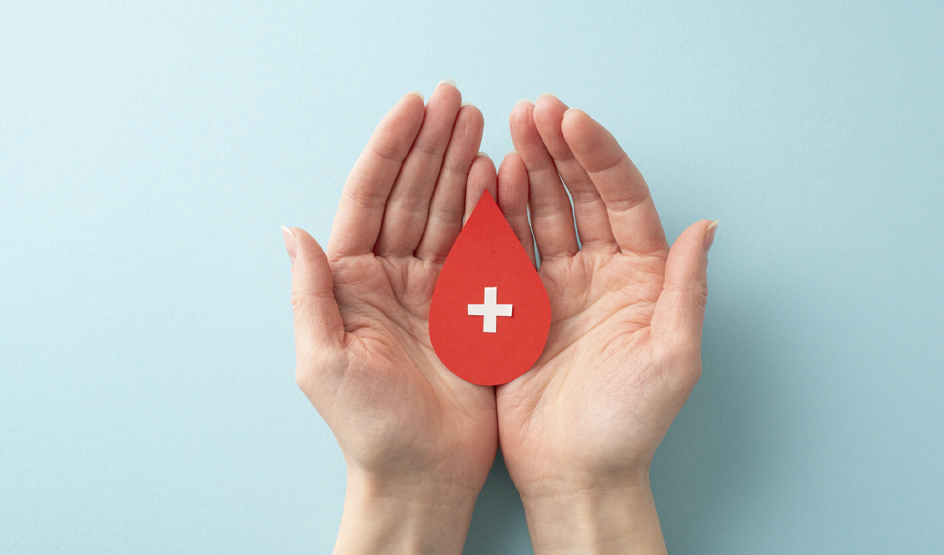 Two hands gently cradle a red blood drop featuring a white cross, symbolizing health and hope.
