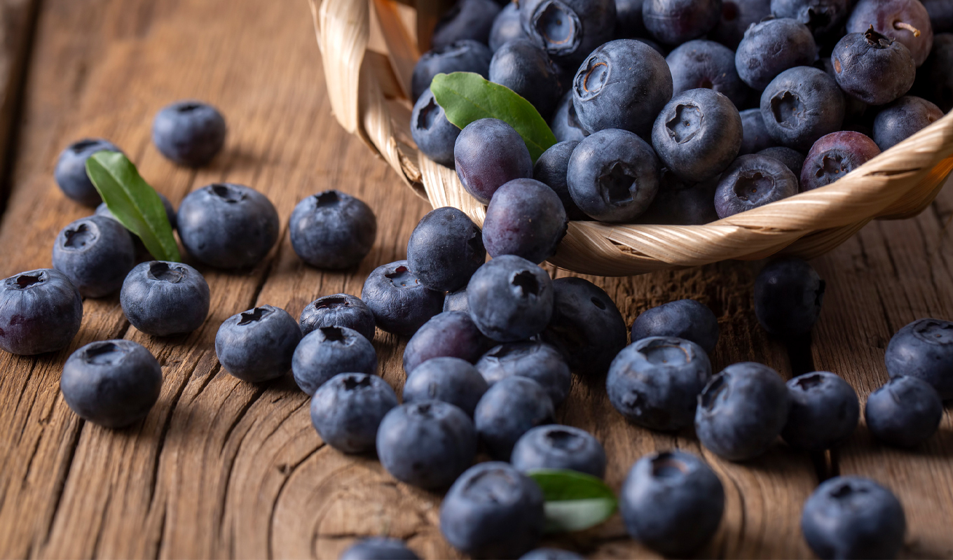 Fresh ripe blueberries spilling from a small woven basket onto a wooden surface, with scattered berries and a few green leaves highlighting natural texture and freshness.