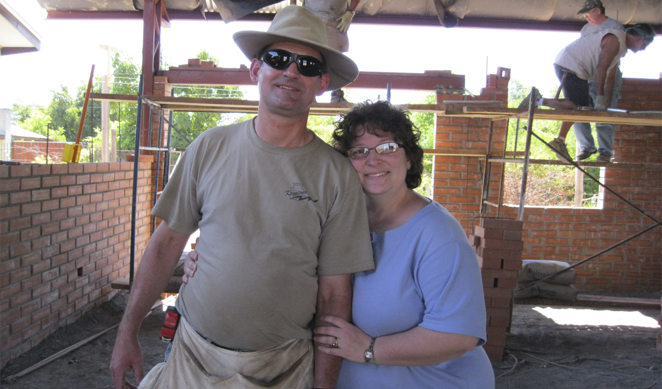 A smiling couple stands close together in front of a brick wall under construction. The man wears a hat, sunglasses, and a khaki t-shirt. The woman, with glasses, rests her hand on his arm. In the background, building laborers stand on scaffolding and continue laying bricks for the wall. Construction materials and tools are scattered around the site.