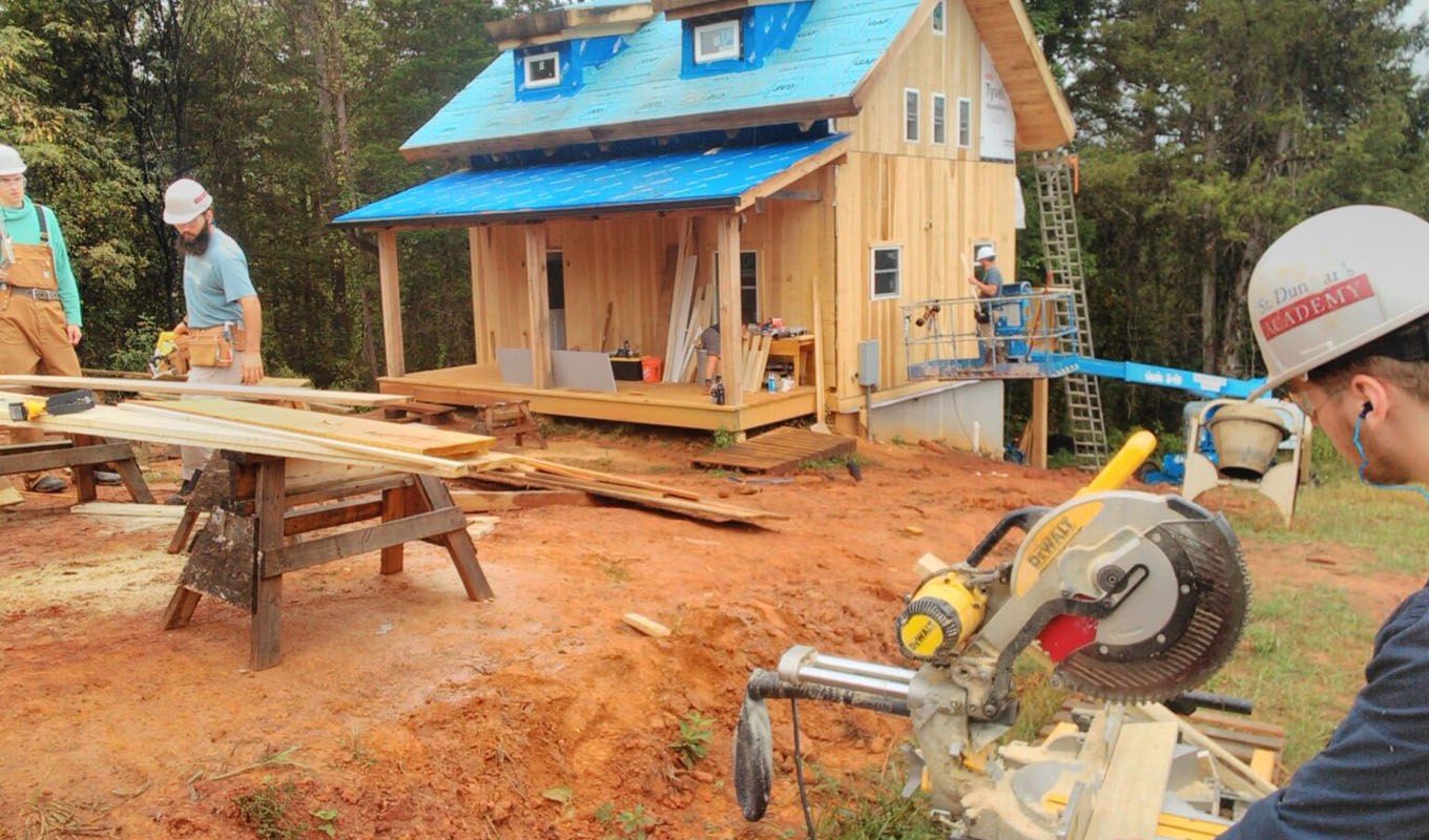 Construction workers build a small wooden house in a wooded area, with lumber and power tools in the foreground.