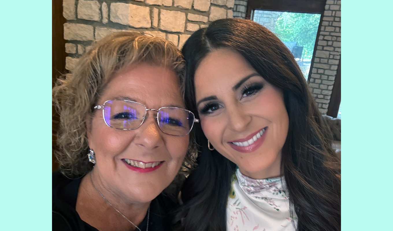 Two people standing close together indoors in front of a stone wall, with one wearing a dark top and the other wearing a light floral-patterned top. There is a window in the background showing greenery outside.