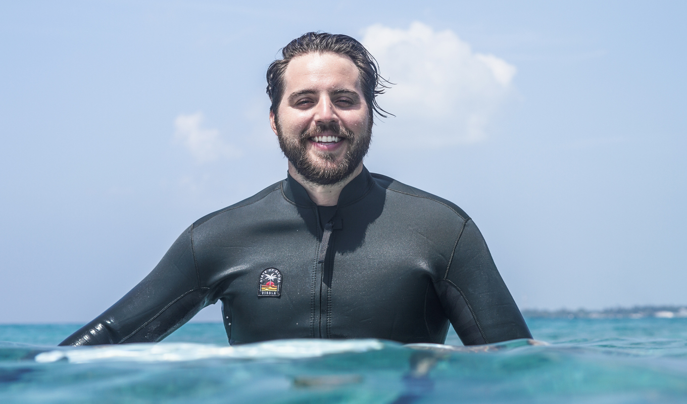 Person in a black wetsuit stands chest-deep in ocean water facing the camera, with blue sky and clouds behind.