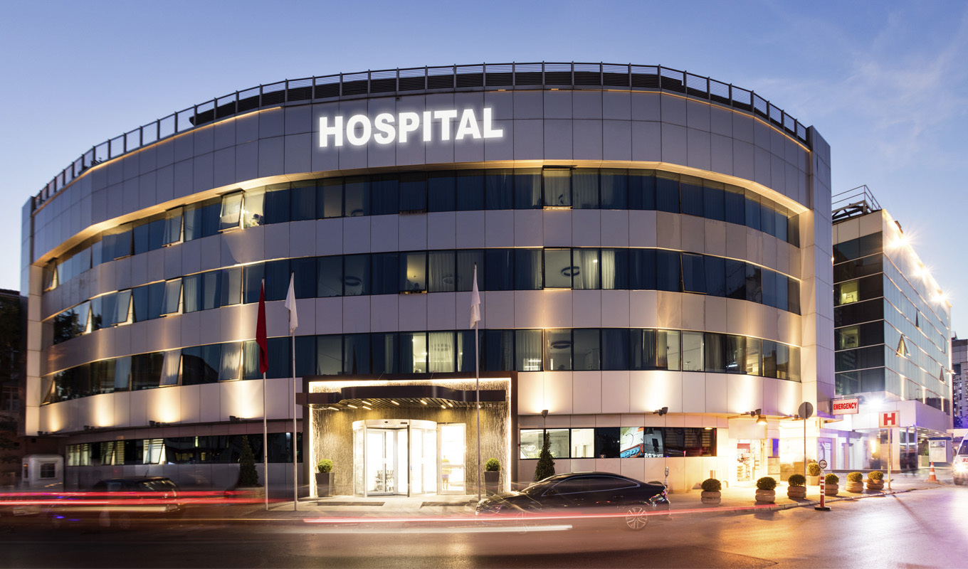 Modern hospital building at dusk with "HOSPITAL" sign and emergency entrance. Streaks of car lights illuminate the street.