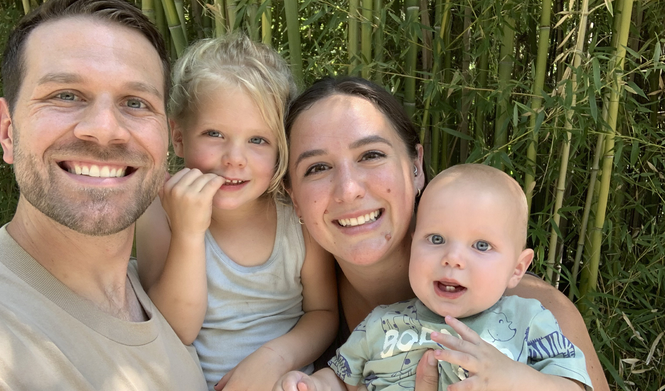 A smiling family consisting of a dad, a mom, a toddler, and an infant stand together in a wooded area.