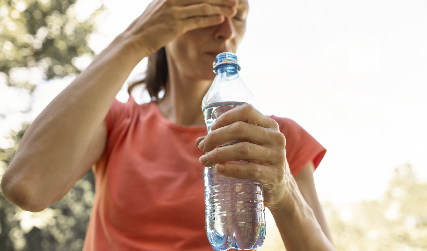 A woman outdoors, wearing a pink t-shirt, holds a water bottle in one hand and shields her eyes with the other, seemingly affected by the heat. Background shows a blurred outdoor environment.