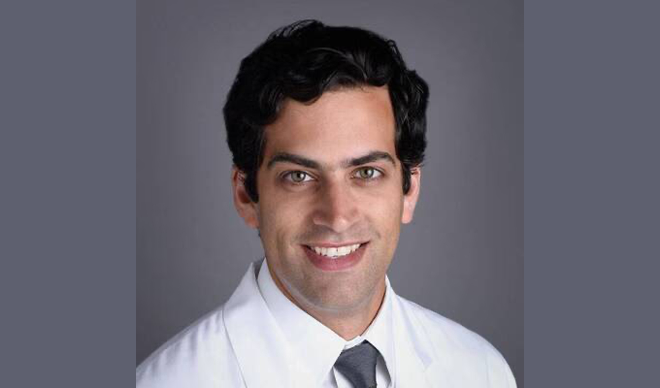 A person wearing a white medical coat and a neatly tied gray necktie is posed against a plain gray background. The individual has dark, wavy hair, and the portrait is centered and professionally lit.