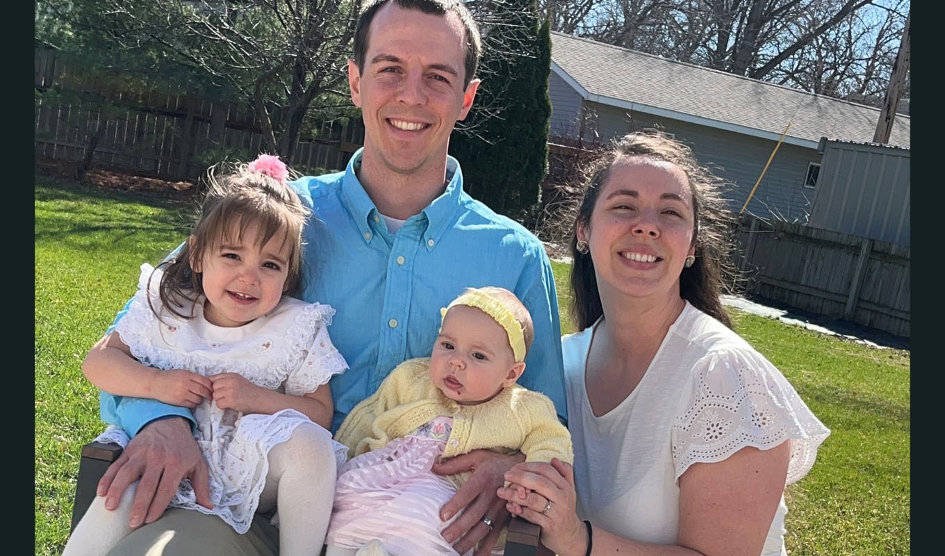 Four people pose outdoors on a sunny day in a grassy backyard. Two adults stand holding two young children.