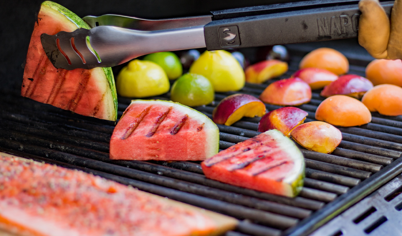 Grilled summer fruit on barbecue grill including watermelon slices, peaches, limes, and lemons with visible grill marks, being turned with metal tongs