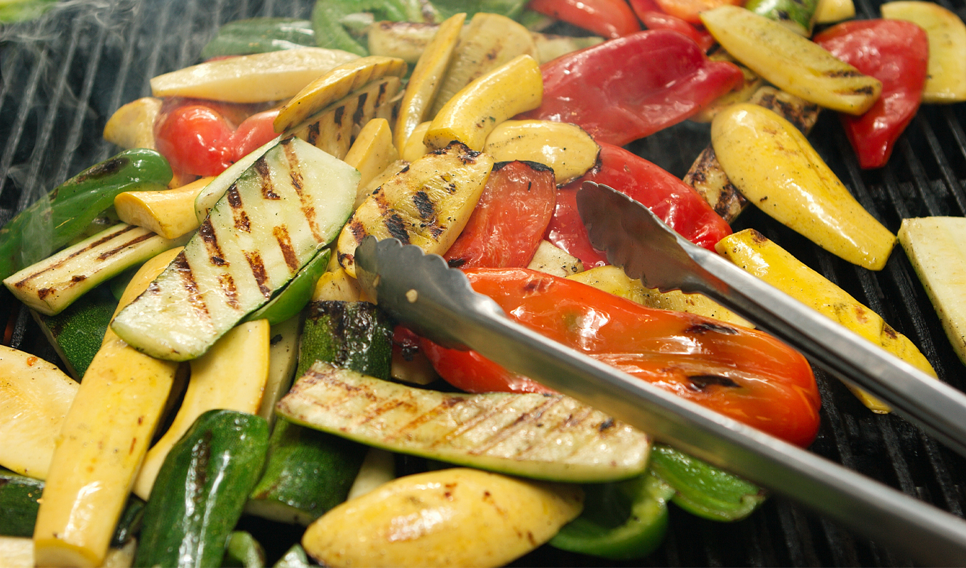 Close-up of mixed grilled vegetables—zucchini slices, yellow squash, and colorful bell peppers—cooking on a barbecue grill with visible grill marks and stainless steel tongs for summer BBQ recipes