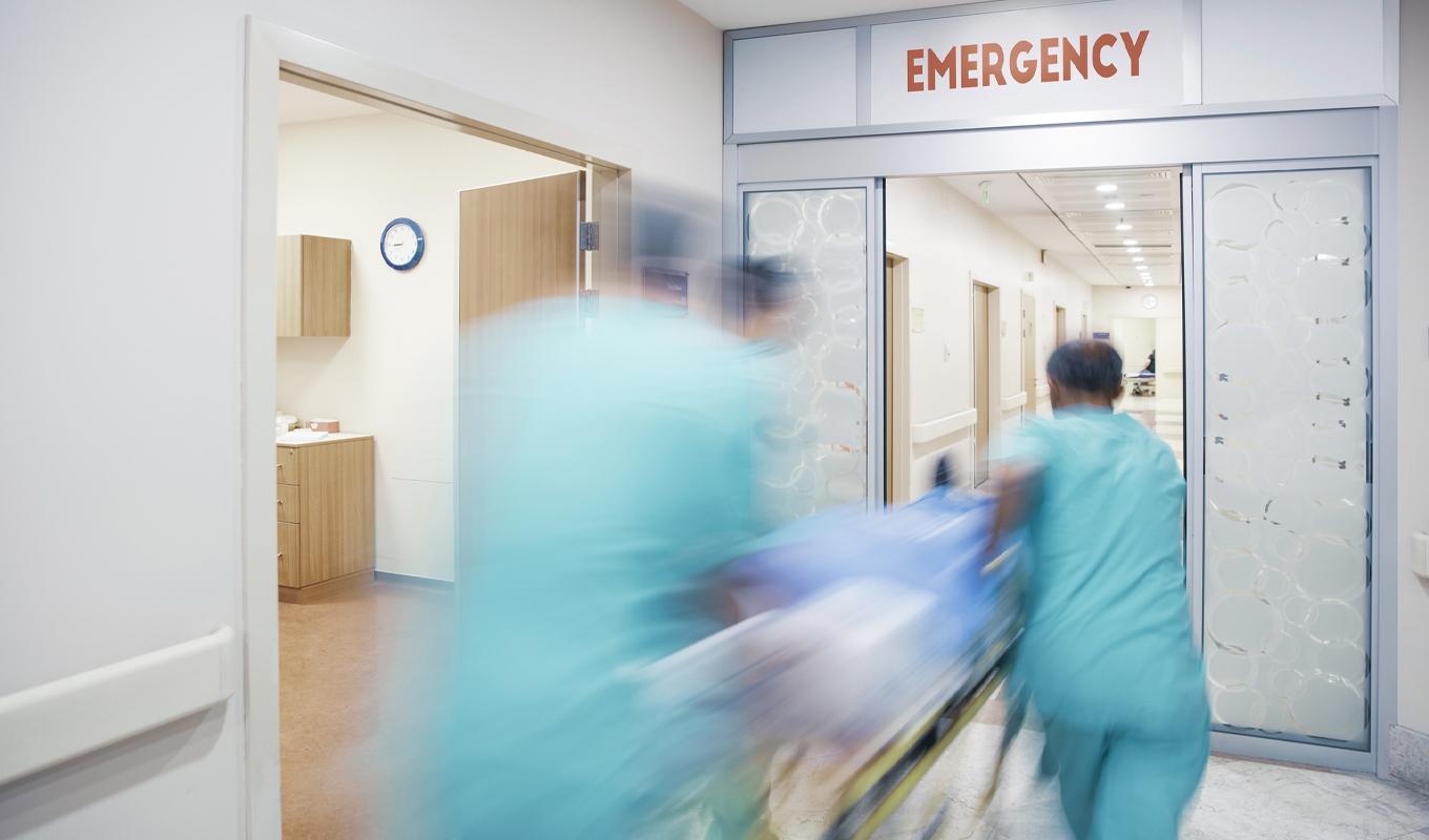 In a brightly lit hospital setting, two medical personnel, wearing teal scrubs, are seen in blurred motion as they urgently push a gurney through automatic sliding doors marked with a red "EMERGENCY" sign. The gurney is covered with a blue sheet. The hallway beyond the doors reveals other doors lining the corridor. To the left, a partially visible hospital room reveals a clock on the wall and cabinets. The overall scene conveys a sense of urgency and medical action.