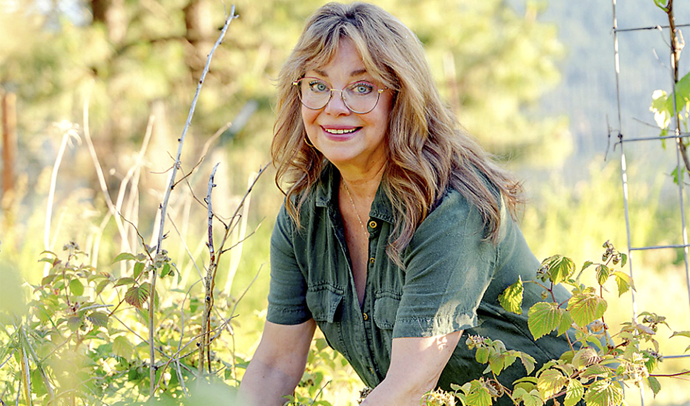 Person gardening among lush green plants under a sunny backdrop, smiling towards the camera.
