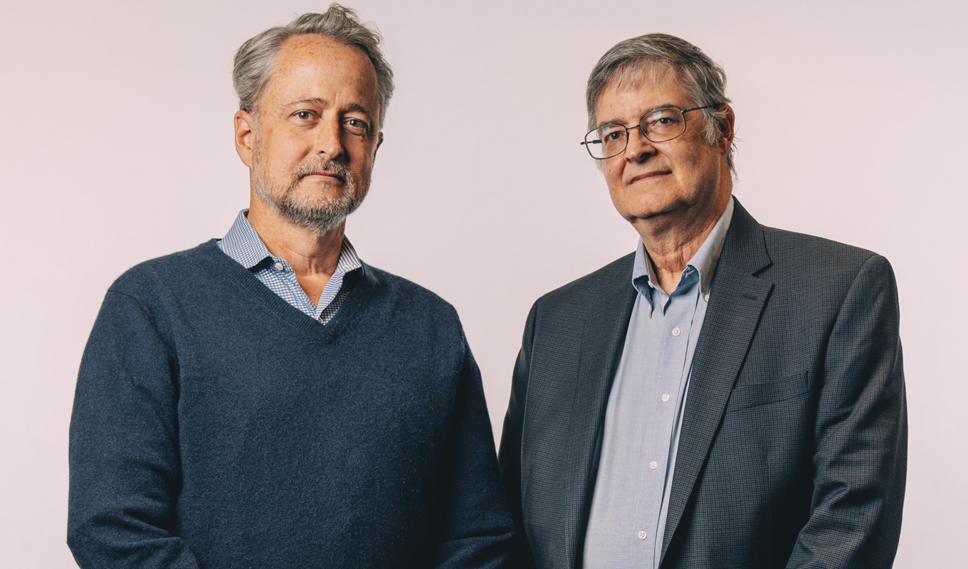 Two men standing side by side in a studio, engaged in conversation, with a neutral background and professional lighting.