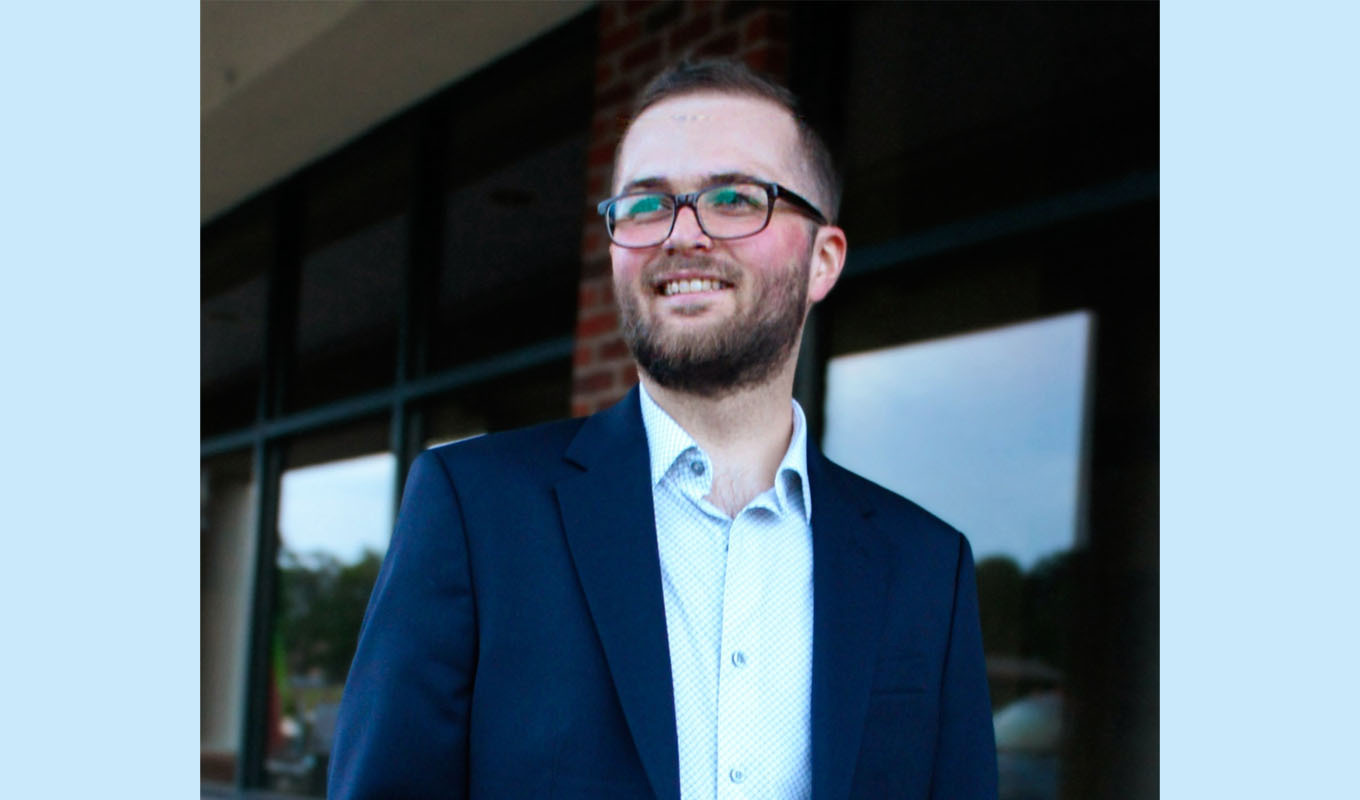 Person wearing glasses and a suit stands outdoors in front of a brick building with large windows, looking slightly to the side.