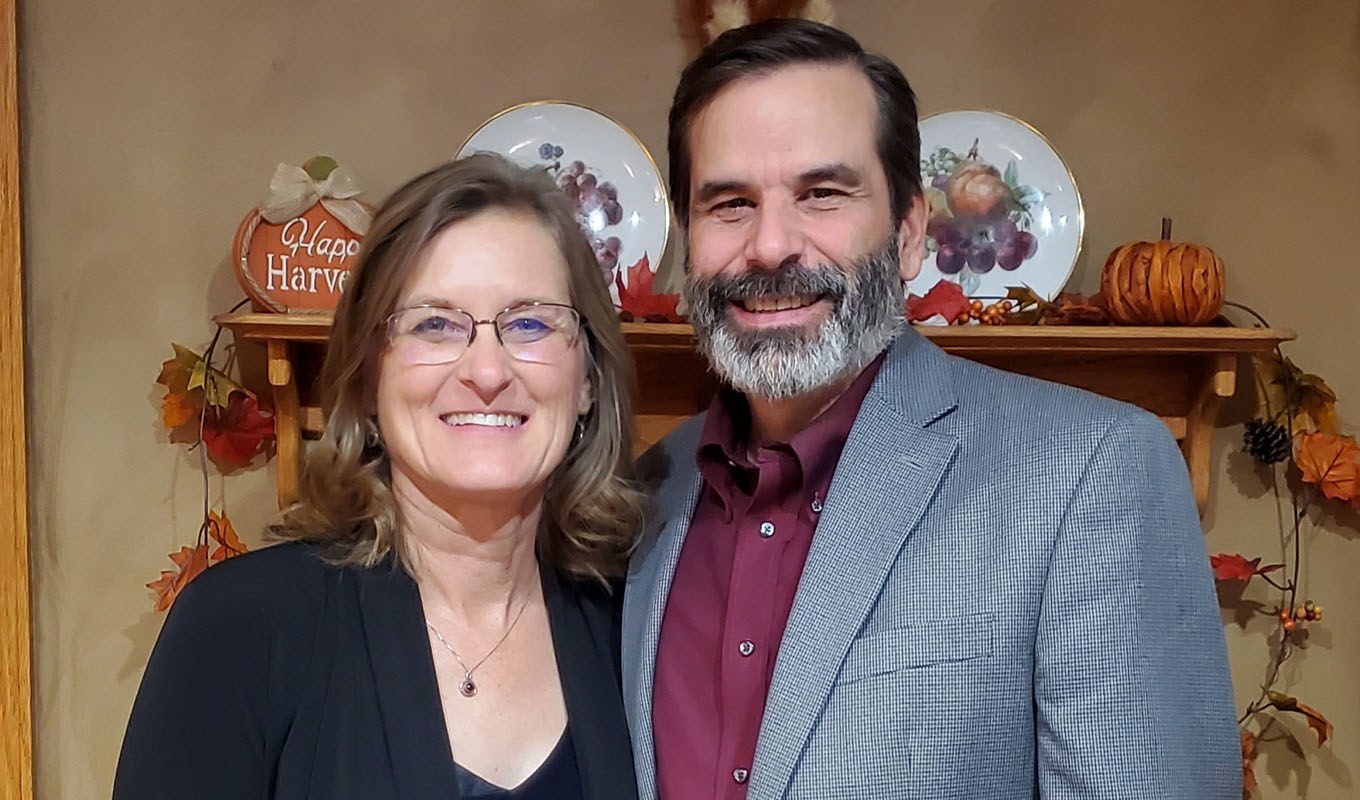 A woman and a man stand side by side indoors, with autumn-themed decorations and plates on a shelf behind them.