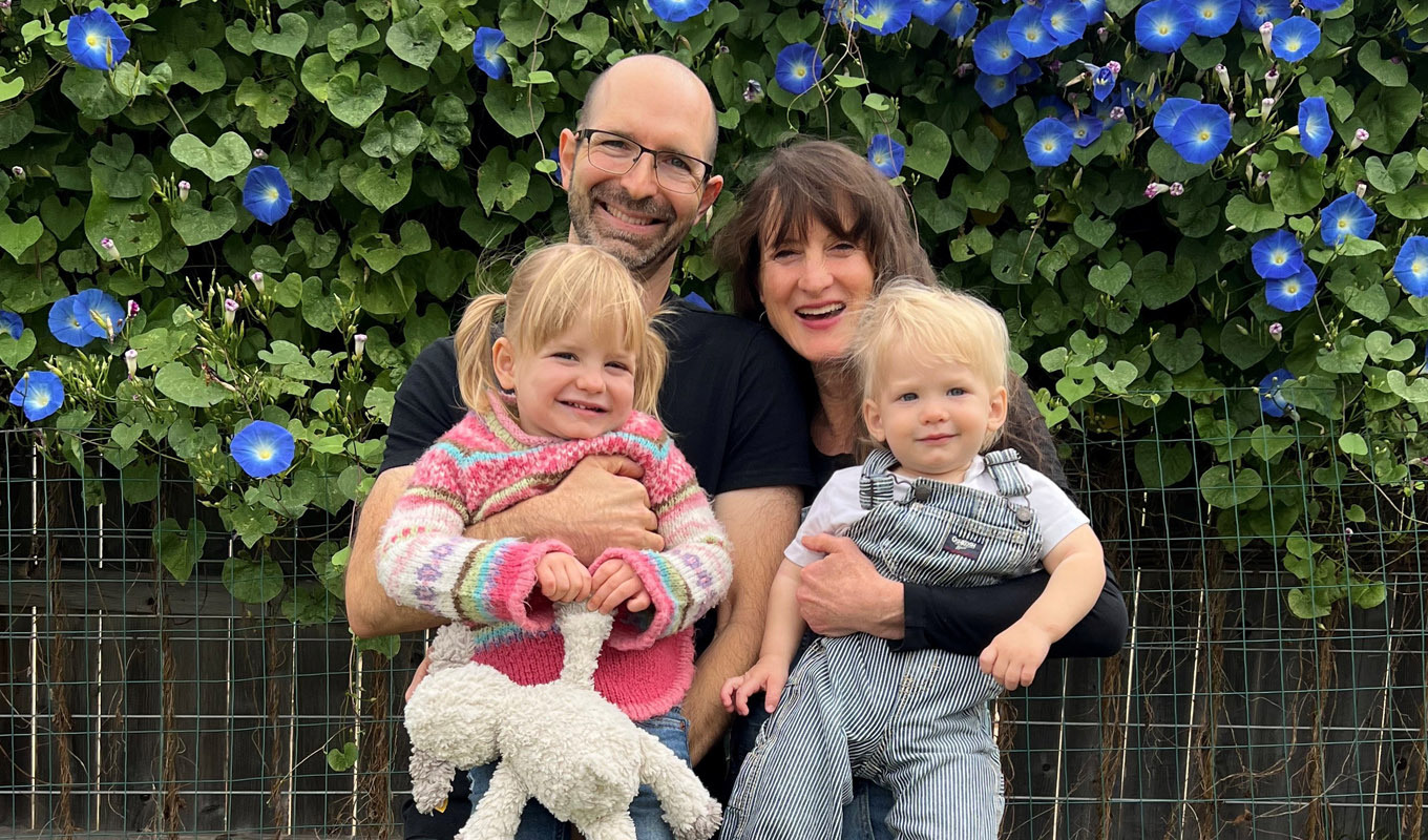 Two adults holding two small children pose in front of leafy vines with bright blue flowers, with a garden fence along the bottom.