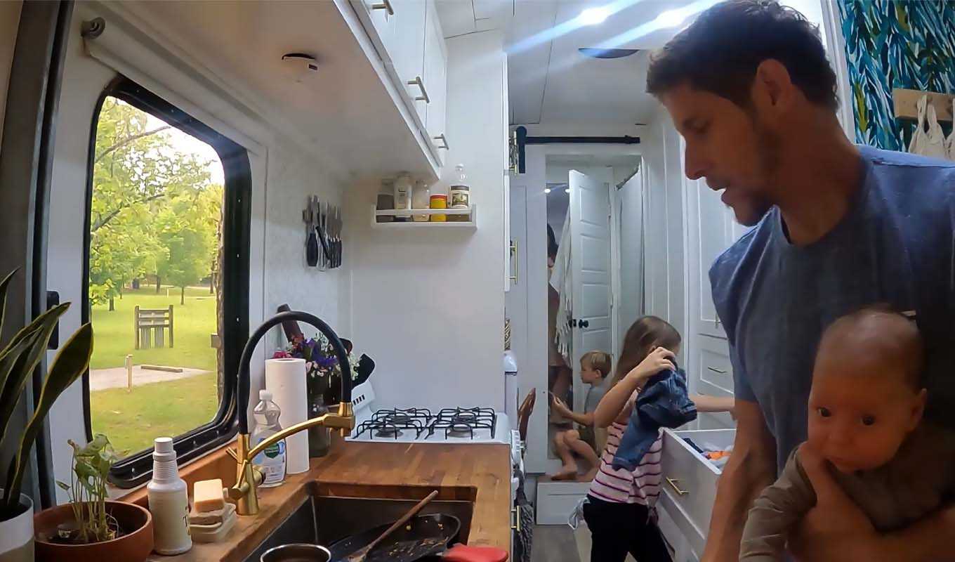 JD Lott holds on to newborn Aquila while other Lott children handle chores in their 200-square-foot bus/home. (YouTube screenshot)