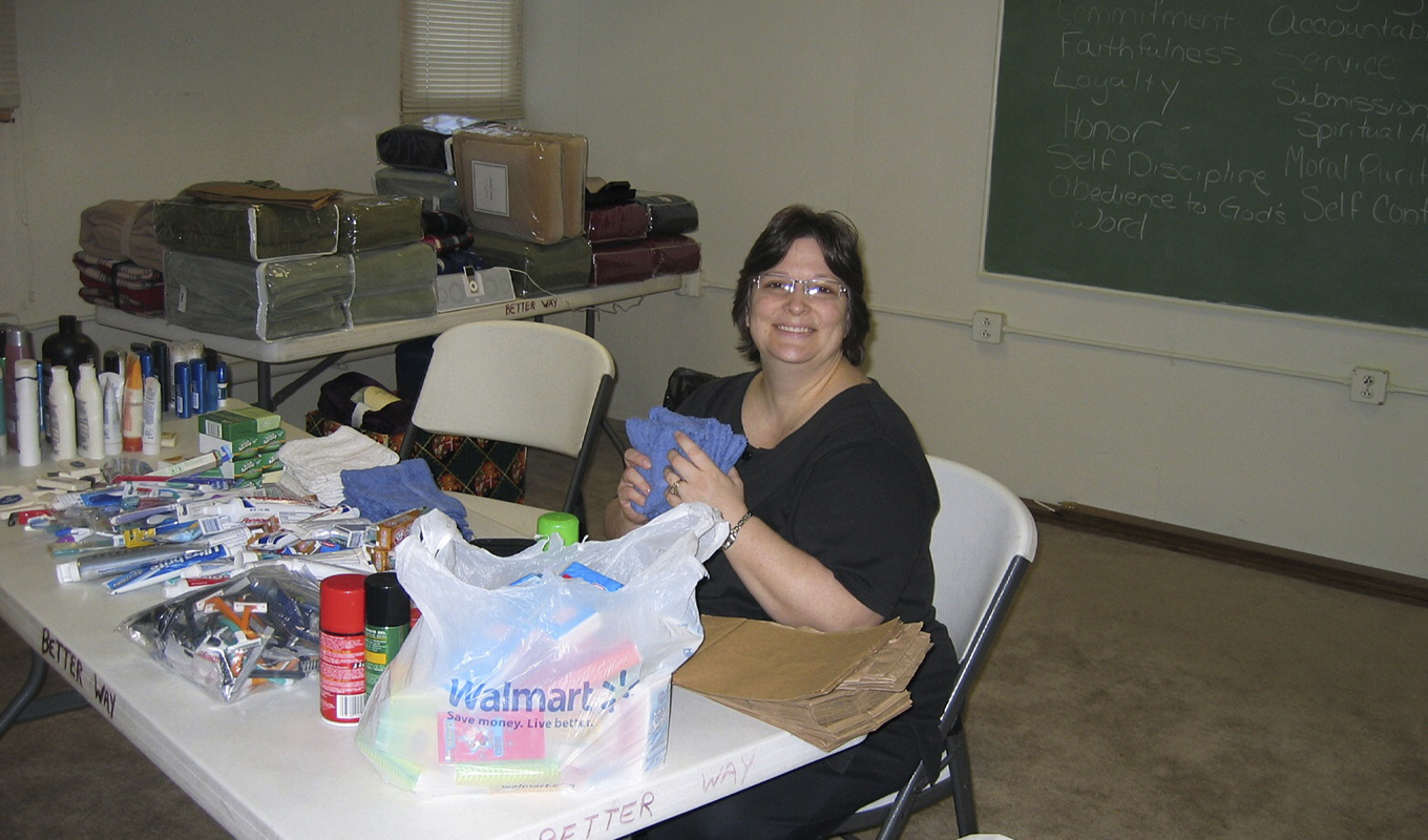 A smiling woman with short brown hair and glasses sits at a table filled with toiletries, linens, and supplies being sorted for the Lydia's Grace program. The table is covered with items such as toothpaste, shampoo, shaving cream, and washcloths. A Walmart bag sits in front of her, ready to be filled with the sorted items. In the background there is a chalkboard with motivational words.