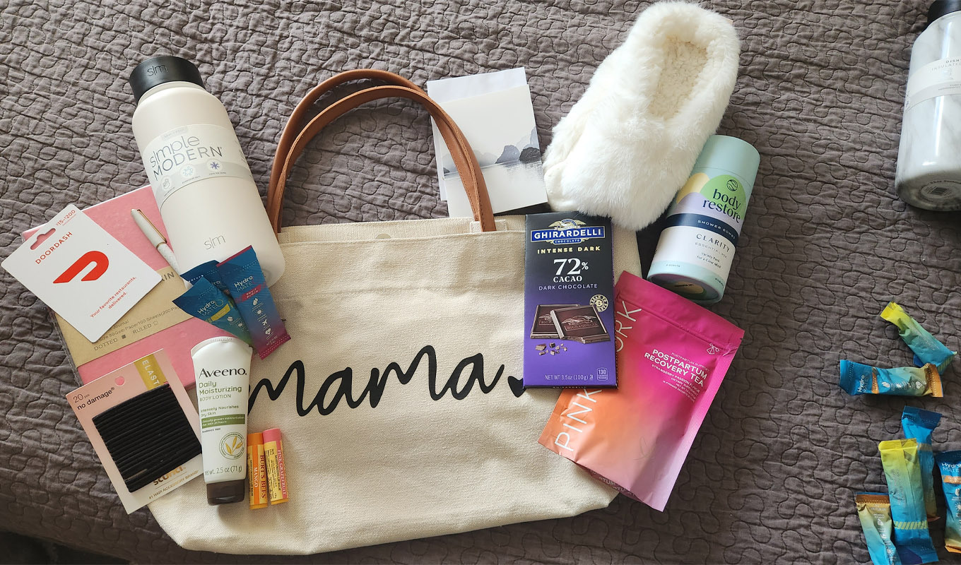 Canvas tote bag labeled “mama” surrounded by comfort items including slippers, chocolate, tea, toiletries, a water bottle, and gift cards, arranged on a bed.