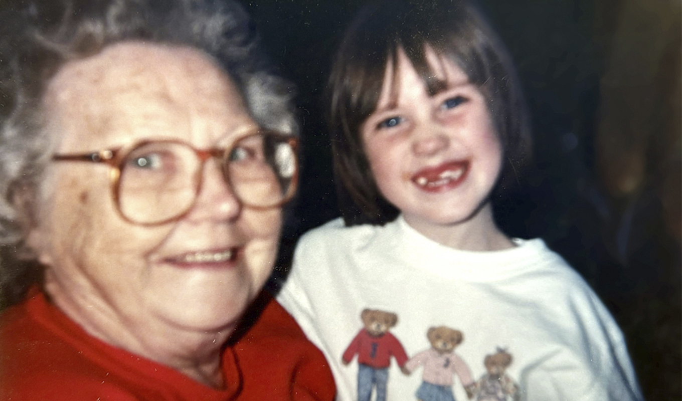 Photograph of an elderly individual and a young child smiling at the camera. The elder is wearing glasses and a red shirt, while the child is in a white shirt featuring cartoon characters.