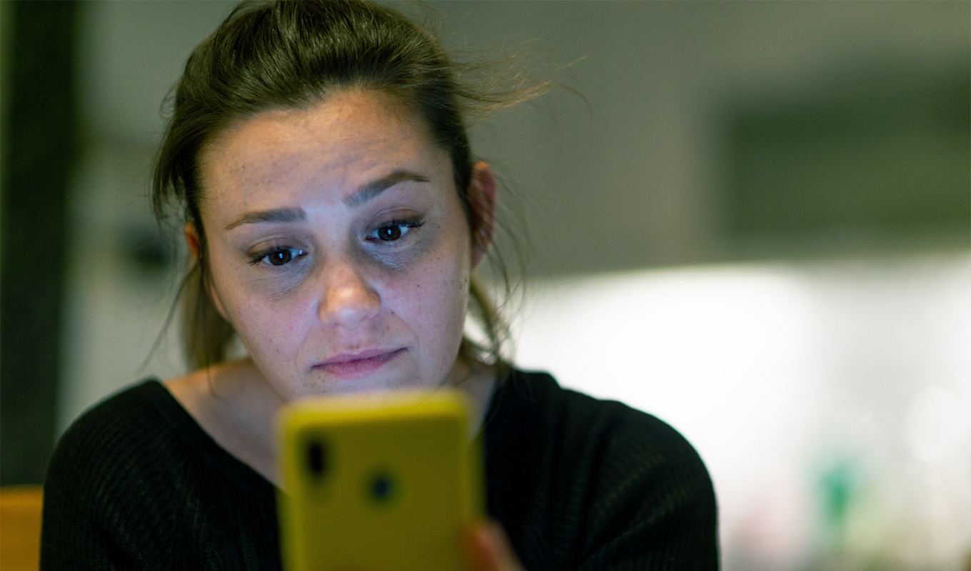 A person with brown hair tied back is holding a bright yellow smartphone in front of them. The individual is wearing a dark-colored top, and the background appears softly blurred, suggesting an indoor setting with neutral tones and diffused lighting.