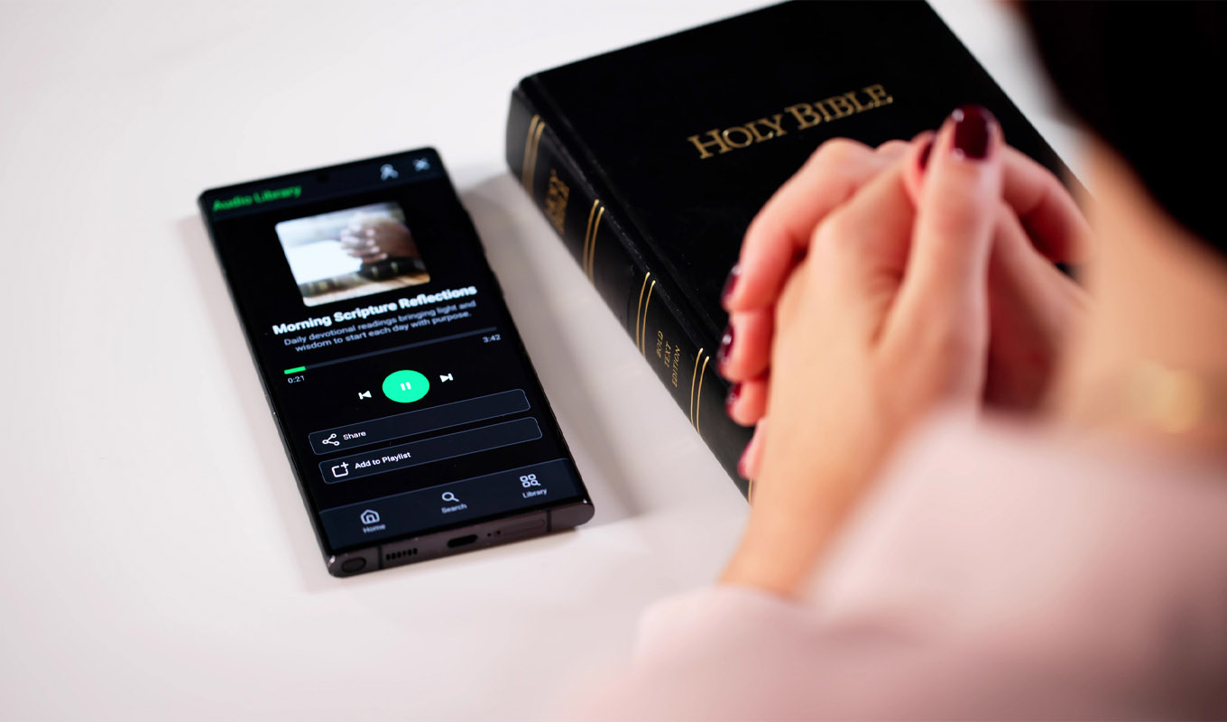 Hands clasped in prayer beside a Holy Bible and a smartphone displaying a devotional audio app on a clean surface.